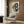 May include: Abstract modern art print with geometric shapes in mustard yellow, navy blue, gray and black tones, displayed in light wood frame on textured wood panel wall. Cream upholstered chair with geometric patterned pillow and brass legs positioned below artwork. Brass floor lamp and dark round side table with open book complete the contemporary reading nook scene.