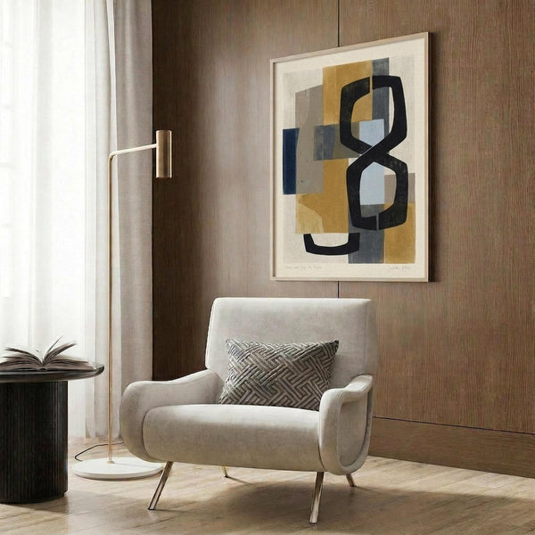 May include: Abstract modern art print with geometric shapes in mustard yellow, navy blue, gray and black tones, displayed in light wood frame on textured wood panel wall. Cream upholstered chair with geometric patterned pillow and brass legs positioned below artwork. Brass floor lamp and dark round side table with open book complete the contemporary reading nook scene.
