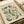May include: Vintage-style botanical art print with decorative floral design featuring green leafy vines, colorful flowers in blue, orange and pink tones, and small berries. The illustration has an ornate, folk art aesthetic with flowing organic curves on cream-colored paper. Print is shown matted in white frame leaning against neutral colored books or papers in soft natural lighting.