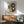 May include: Abstract art print in wooden frame hanging above mid-century modern walnut credenza with white ceramic table lamp. Artwork features geometric shapes in navy blue, mustard yellow, gray and cream tones. Green upholstered armchair with wooden arms positioned nearby. Bright natural sunlight streams across white wall creating dramatic shadows in minimalist living room setting.