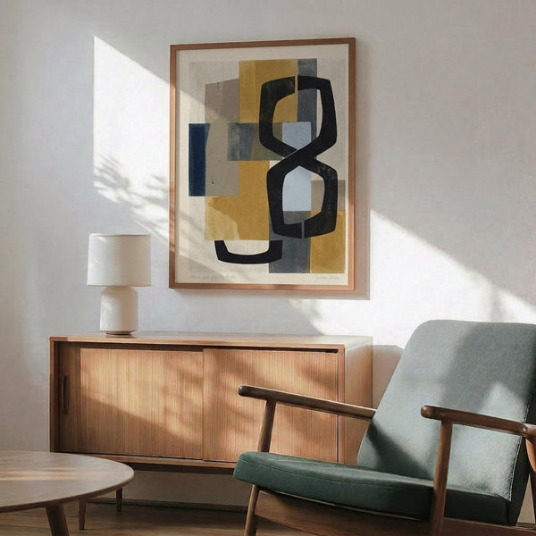May include: Abstract art print in wooden frame hanging above mid-century modern walnut credenza with white ceramic table lamp. Artwork features geometric shapes in navy blue, mustard yellow, gray and cream tones. Green upholstered armchair with wooden arms positioned nearby. Bright natural sunlight streams across white wall creating dramatic shadows in minimalist living room setting.