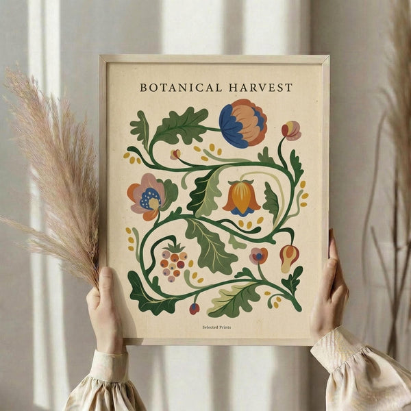 May include: Hands holding a framed botanical art print featuring decorative folk-style flowers and vines in blue, orange, and green on cream background. Light wood frame, neutral beige clothing, dried wheat stalks visible, soft natural lighting against white curtained window.
