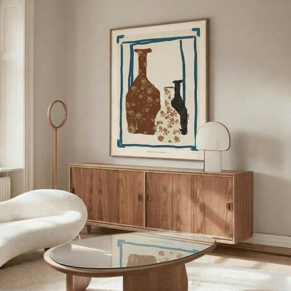 May include: Modern living room with walnut sideboard displaying framed abstract art print featuring three vessels in brown, white, and black with blue border on cream background. Round glass coffee table reflects the artwork below. White curved chair and brass floor lamp complete the minimalist interior styling.