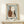 May include: Framed abstract still life print featuring three pottery vessels in rust brown, white with terracotta spots, and dark blue colors, surrounded by hand-painted blue border on cream paper. Light wood frame displayed on white wooden console with small wrapped gift box beside it against cream wall.