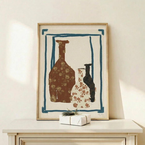 May include: Framed abstract still life print featuring three pottery vessels in rust brown, white with terracotta spots, and dark blue colors, surrounded by hand-painted blue border on cream paper. Light wood frame displayed on white wooden console with small wrapped gift box beside it against cream wall.