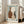 May include: Framed art print leaning against white wall next to beige upholstered chair in bright living room with large windows. Print features three pottery vessels in earthy brown, cream with rust spots, and dark teal, surrounded by hand-drawn blue border on cream background. Light wood frame complements neutral room decor and hardwood floors.