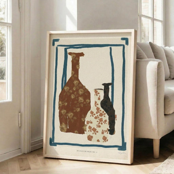 May include: Framed art print leaning against white wall next to beige upholstered chair in bright living room with large windows. Print features three pottery vessels in earthy brown, cream with rust spots, and dark teal, surrounded by hand-drawn blue border on cream background. Light wood frame complements neutral room decor and hardwood floors.