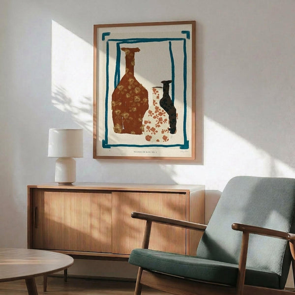 May include: Framed art print displaying three ceramic vessels in brown, white with red spots, and dark colors against cream background with teal border, shown mounted on white wall above wooden sideboard with modern green upholstered chair and table lamp in minimalist room setting with natural lighting creating geometric shadows.