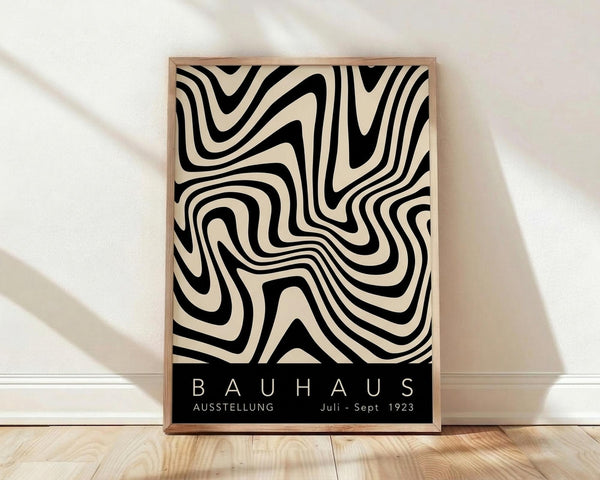 May include: Framed Bauhaus exhibition poster with black and cream swirled optical illusion pattern leaning against white wall on light wood floor. Natural shadows from window light create diagonal patterns across white fabric backdrop. Wooden frame has natural grain visible around geometric artwork featuring flowing, hypnotic curves and text reading "BAUHAUS AUSSTELLUNG Juli-Sept 1923" in white letters on black band at bottom.