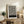 May include: Black and white abstract poster with flowing curved lines in a wooden frame, displayed on a rustic wooden sideboard next to a fern plant, ceramic vase, and stacked books in a neutral-toned living room setting with beige furniture and soft natural lighting.