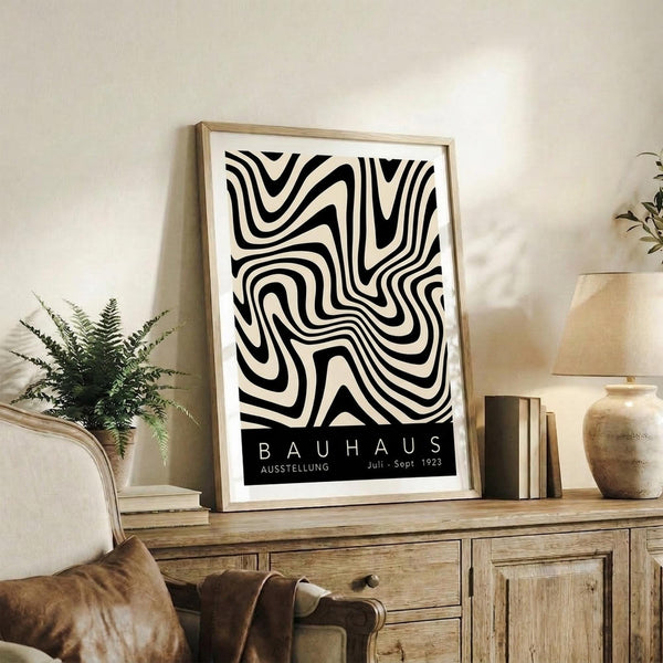 May include: Black and white abstract poster with flowing curved lines in a wooden frame, displayed on a rustic wooden sideboard next to a fern plant, ceramic vase, and stacked books in a neutral-toned living room setting with beige furniture and soft natural lighting.