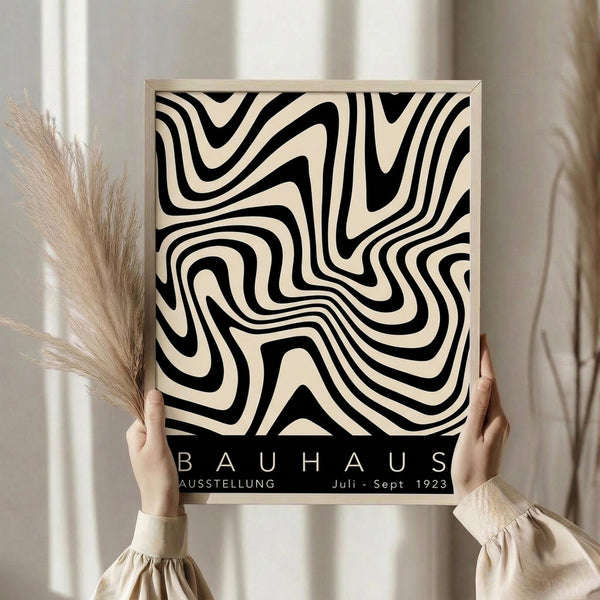 May include: Person in cream ruffled sleeves holding framed black and white abstract swirling pattern poster against white curtained window, with dried pampas grass visible to the left. The artwork features flowing organic lines creating a mesmerizing optical illusion effect, with "BAUHAUS AUSSTELLUNG Juli-Sept 1923" text at bottom in minimalist typography.