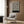May include: Black and white swirling abstract pattern poster with "BAUHAUS AUSSTELLUNG Juli-Sept 1923" text mounted on wood-paneled wall above beige upholstered armchair with geometric patterned pillow. Modern brass floor lamp with white circular base stands nearby next to dark round side table with open book. Neutral-toned room setting with curtained window and wooden flooring.
