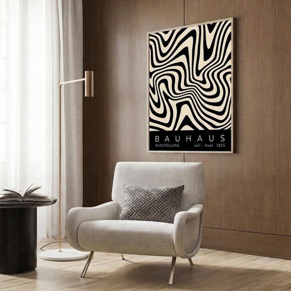 May include: Black and white swirling abstract pattern poster with "BAUHAUS AUSSTELLUNG Juli-Sept 1923" text mounted on wood-paneled wall above beige upholstered armchair with geometric patterned pillow. Modern brass floor lamp with white circular base stands nearby next to dark round side table with open book. Neutral-toned room setting with curtained window and wooden flooring.