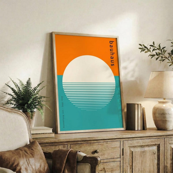 May include: Framed Bauhaus-style poster with orange and teal geometric design featuring white circle and horizontal lines, displayed on rustic wooden sideboard alongside fern plant, books, ceramic vase, and brown leather chair in modern living room setting with white walls