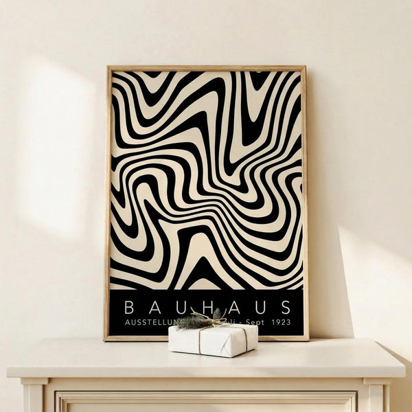 May include: A framed black and cream poster featuring bold, flowing abstract patterns reminiscent of zebra stripes or topographical lines. The design has organic, wavy curves that create an optical illusion effect. At the bottom is a black band with white text reading "BAUHAUS" and exhibition details from 1923. The poster sits in a thin wooden frame on a light wood surface next to a small white wrapped gift with twine, against a cream-colored wall.