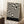 May include: Black and cream swirling abstract pattern poster in light wood frame, leaning against white wall corner. Black text band at bottom reads "BAUHAUS AUSSTELLUNG Juli-Sept 1923". Coffee cup on wooden block and beige knit throw visible nearby. Soft natural lighting creates gentle shadows on textured white rug.