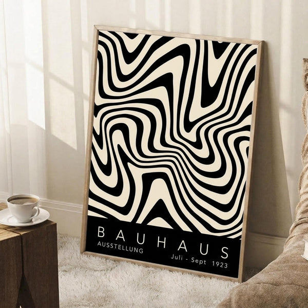 May include: Black and cream swirling abstract pattern poster in light wood frame, leaning against white wall corner. Black text band at bottom reads "BAUHAUS AUSSTELLUNG Juli-Sept 1923". Coffee cup on wooden block and beige knit throw visible nearby. Soft natural lighting creates gentle shadows on textured white rug.