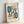 May include: Framed poster with abstract geometric design featuring teal oval shapes, orange vertical elements, black angular lines, circles, and rectangular patterns with concentric lines. Text reads "BAUHAUS" at top and "Ausstellung Juli - Sept 1923" at bottom. Light wood frame against white wall, casting soft shadows.