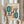 May include: Person in beige sweater holding framed Bauhaus exhibition poster featuring geometric shapes in teal blue and orange with black linear elements and concentric rectangles on cream background. Dried pampas grass visible to left against white curtained window. Light wood frame, minimalist modern interior setting.