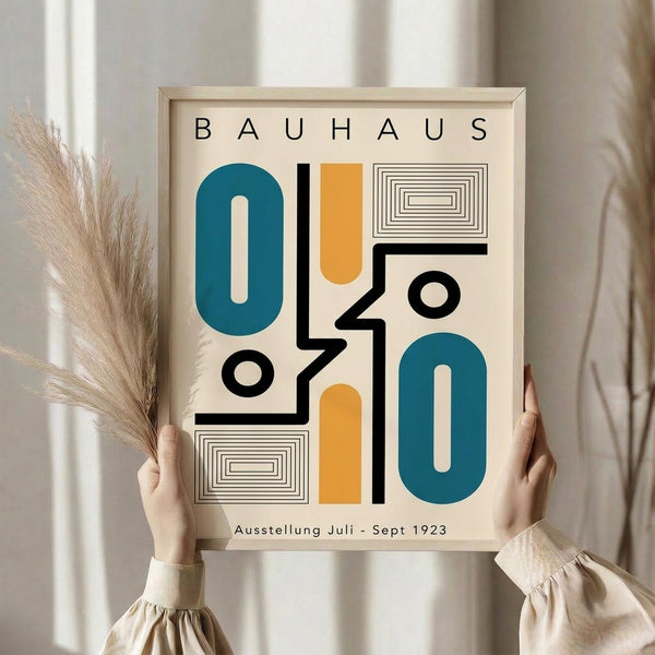 May include: Person in beige sweater holding framed Bauhaus exhibition poster featuring geometric shapes in teal blue and orange with black linear elements and concentric rectangles on cream background. Dried pampas grass visible to left against white curtained window. Light wood frame, minimalist modern interior setting.