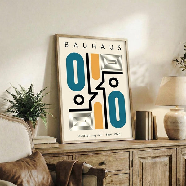 May include: Framed Bauhaus-style poster with geometric shapes in teal, orange, and black displayed on wooden sideboard next to fern plant, books, and ceramic vase in neutral living room setting with beige armchair visible