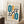 May include: Framed Bauhaus-style poster with geometric abstract design featuring teal ovals, orange rounded rectangles, black lines and circles, and concentric rectangle patterns on cream background. Light wood frame leans against white wall corner next to wooden block with white coffee cup and saucer, on textured white rug with beige blanket visible.