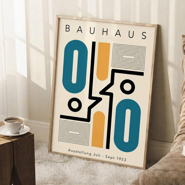 May include: Framed Bauhaus-style poster with geometric abstract design featuring teal ovals, orange rounded rectangles, black lines and circles, and concentric rectangle patterns on cream background. Light wood frame leans against white wall corner next to wooden block with white coffee cup and saucer, on textured white rug with beige blanket visible.
