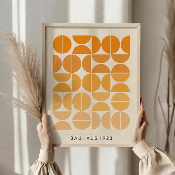 May include: Person in beige clothing holding framed geometric art print featuring orange and yellow circles and semicircles in grid pattern on cream background, with dried pampas grass stems, against soft neutral curtained backdrop