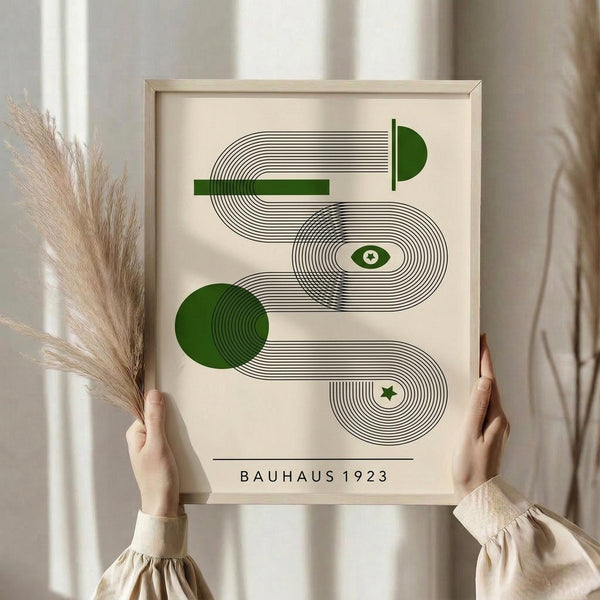 May include: Person in beige clothing holding framed geometric abstract print with green circles, curved black lines, eye and star symbols in cream-colored frame. Dried pampas grass held in other hand against soft neutral background with natural window light.