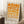 May include: Framed geometric art print with orange and cream circular pattern in light wood frame, leaning against white wall corner next to white coffee cup on wooden block and beige textured throw blanket on light colored rug