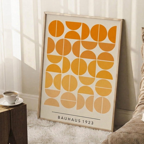 May include: Framed geometric art print with orange and cream circular pattern in light wood frame, leaning against white wall corner next to white coffee cup on wooden block and beige textured throw blanket on light colored rug