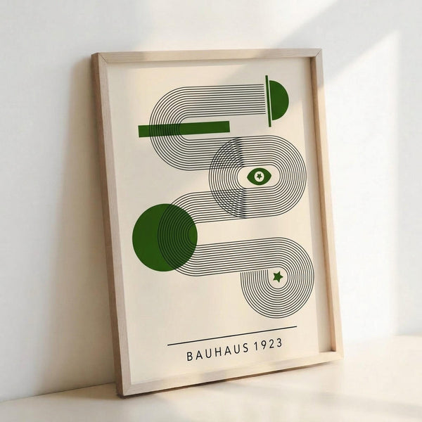 Framed geometric art print featuring flowing curved lines in black ink with green accents including circles, rectangles, an eye symbol, and star. The minimalist design uses concentric line patterns on cream paper with "BAUHAUS 1923" text at bottom. Natural wood frame leans against white wall, showing warm lighting and shadows.