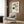 May include: Cream poster with green and black geometric Bauhaus design featuring curved lines, circles, and an eye motif, displayed in light wood frame on wooden wall panel. Scene shows beige upholstered armchair with patterned pillow, brass floor lamp with white base, and dark round side table with open book. Natural light from window with curtains creates warm, modern interior setting.