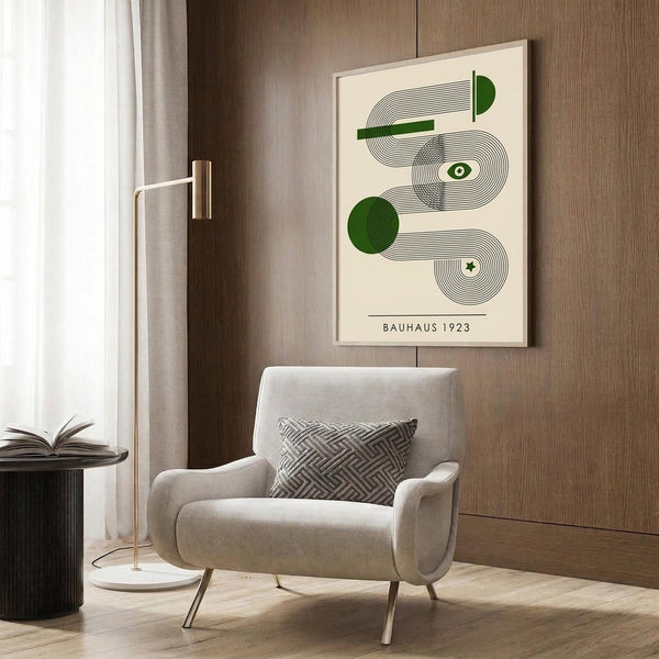May include: Cream poster with green and black geometric Bauhaus design featuring curved lines, circles, and an eye motif, displayed in light wood frame on wooden wall panel. Scene shows beige upholstered armchair with patterned pillow, brass floor lamp with white base, and dark round side table with open book. Natural light from window with curtains creates warm, modern interior setting.