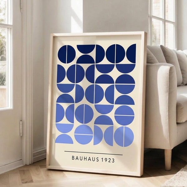 May include: Framed geometric poster with blue circles and semicircles arranged in a grid pattern on cream background, displayed in bright living room next to beige sofa. Natural wood frame, sunlight streaming through white windows onto hardwood floors.