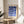 May include: Framed geometric poster with blue circles and semicircles arranged in a grid pattern on cream background, displayed on light wood round side table next to beige upholstered chair. Natural lighting creates soft shadows across neutral-toned room with plant visible in corner.