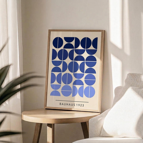 May include: Framed geometric poster with blue circles and semicircles arranged in a grid pattern on cream background, displayed on light wood round side table next to beige upholstered chair. Natural lighting creates soft shadows across neutral-toned room with plant visible in corner.