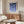 May include: A framed poster with blue geometric circles in various shades arranged in a grid pattern on cream background, displayed above a mid-century modern wooden sideboard. The minimalist room features a white curved chair, glass-top coffee table, brass floor lamp, and neutral walls with natural lighting creating soft shadows.