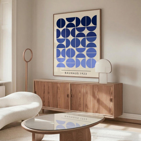 May include: A framed poster with blue geometric circles in various shades arranged in a grid pattern on cream background, displayed above a mid-century modern wooden sideboard. The minimalist room features a white curved chair, glass-top coffee table, brass floor lamp, and neutral walls with natural lighting creating soft shadows.