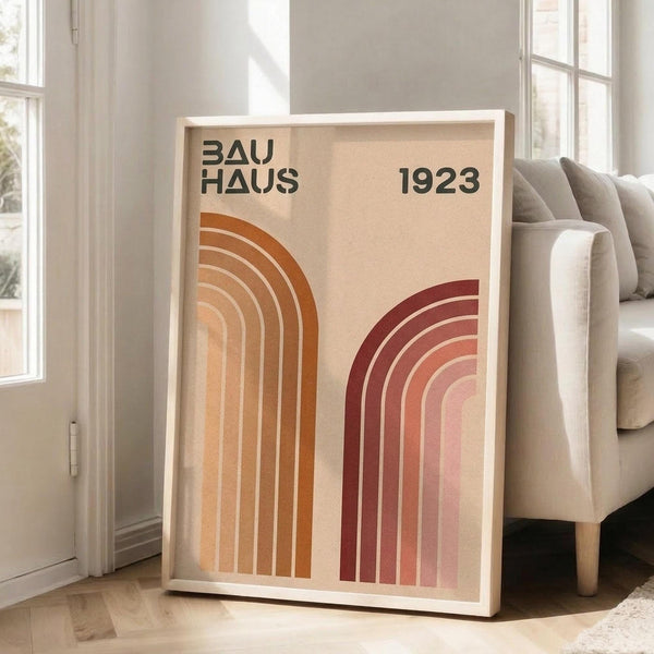 May include: Framed Bauhaus-inspired poster with geometric rainbow arcs in warm orange and burgundy tones against cream background, leaning against white wall next to beige upholstered sofa in bright, minimalist living room with large windows and natural light casting shadows on light wood floors.