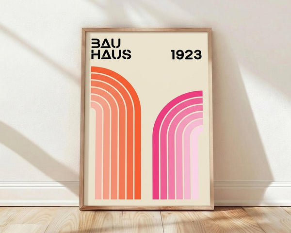 May include: A framed poster with cream background displaying "BAUHAUS" and "1923" in black text at top, featuring two sets of curved concentric lines - orange gradient arcs on left transitioning from deep orange to pale peach, and pink gradient arcs on right transitioning from bright magenta to soft pink. The wooden frame leans against white wall on light wood flooring with natural shadows.