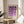 May include: Framed geometric poster with purple circles and semicircles arranged in a grid pattern on cream background, displayed on round wooden side table next to beige upholstered chair. Natural lighting creates soft shadows across neutral-toned room with plant leaves visible in corner.
