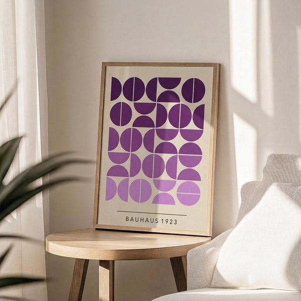 May include: Framed geometric poster with purple circles and semicircles arranged in a grid pattern on cream background, displayed on round wooden side table next to beige upholstered chair. Natural lighting creates soft shadows across neutral-toned room with plant leaves visible in corner.