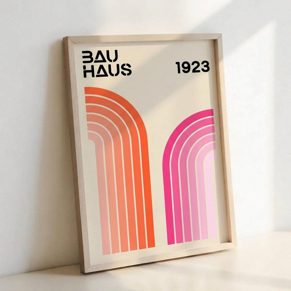 May include: A framed poster with cream background featuring "BAUHAUS 1923" text in black sans-serif letters at top. Two curved geometric shapes create an arch design - left curve transitions from bright orange to pale peach, right curve fades from hot pink to light pink. Both shapes are made of concentric curved lines. The print is displayed in a light wood frame, leaning against a white wall on a neutral surface.