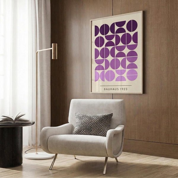 May include: Framed geometric art print featuring purple circles and semicircles in gradient shades from deep violet to light lavender on cream background, displayed above beige upholstered armchair with geometric patterned pillow. Modern reading corner with brass floor lamp, side table with open book, and warm wood paneled walls.