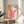 May include: Framed geometric poster with orange and pink curved rainbow arches on cream background, displayed on round wooden side table next to beige upholstered chair. Natural lighting creates soft shadows across white wall. Green plant leaves visible in corner. Modern minimalist home decor styling with neutral tones and clean lines.