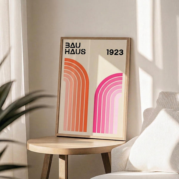 May include: Framed geometric poster with orange and pink curved rainbow arches on cream background, displayed on round wooden side table next to beige upholstered chair. Natural lighting creates soft shadows across white wall. Green plant leaves visible in corner. Modern minimalist home decor styling with neutral tones and clean lines.