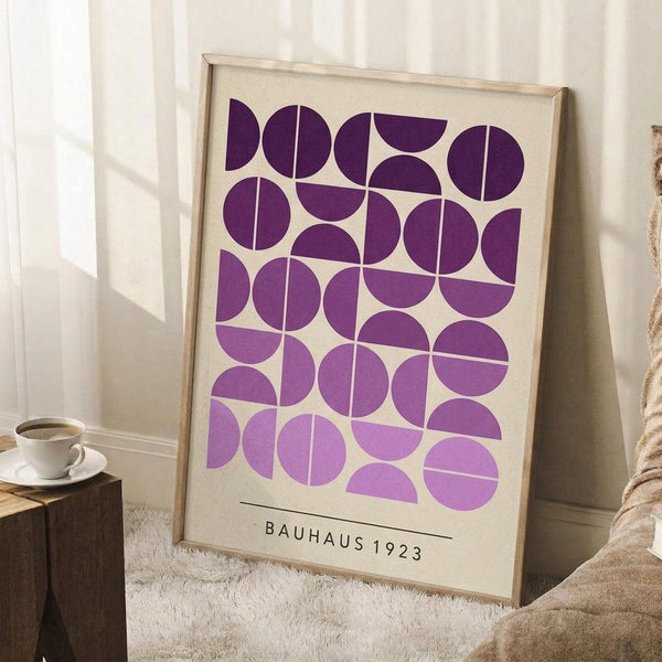 May include: Framed geometric art print with purple circles and semicircles arranged in a grid pattern on cream background, displayed leaning against white wall corner. Print shows "BAUHAUS 1923" text at bottom. Scene includes wooden side table with white coffee cup and saucer, textured cream rug, and soft natural lighting creating shadows on the wall.