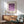 May include: Framed geometric poster with purple and lavender circles and semicircles arranged in a grid pattern on cream background, displayed above mid-century modern wood credenza with ceramic table lamp, next to sage green upholstered armchair with wooden arms in sunlit room with white walls.